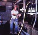 An adult male handles milking equipment while sitting in a swivel chair thatâ€™s attached to the roller track which has been mounted on dairy parlor pit wall.