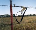 A metal fence-wire post with the Fence Buddy toolâ€™s key componentsâ€”the ratcheting mechanism, the ratchet tie-down strap, and the champ thatâ€™s on the fenceâ€™s topmost wire holding a length of loose wire.