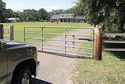 Pictured on a gravel lane leading to a home is a vehicle approaching a wide metal four-rail gate thatâ€™s in the process of opening, the mounting arms attached to gate and hinged gate post.