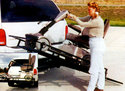 Hitched behind a vehicle is the carrier with a scooter that an adult female has guided onto the tilted platform.  A small inset shows the platform with scooter parallel to the ground.