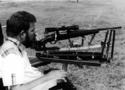 An adult male in s wheelchair hand-control-operating (as described) and firing (via sip-and-puff) the shortened-stock rifle, its barrel resting on the deviceâ€™s T-post.