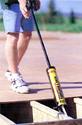 An adult male (standing upright) is using the device to apply adhesive to the top of a floor joist in preparation for another sheet of subflooring.