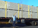 Man with back to camera in blue baseball cap light blue shirt & dark blue pants holding long telescoping pole with attachment on upper end holding a rolled-up cargo strap that he is throwing over the side of a semi trailer