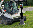 A loader-mounted fence-wire winder mounted on a white skid steer sitting outside in the grass.