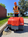 Man in orange hardhat and reflective vest walking behind a heavy-duty walk-behind sweeper